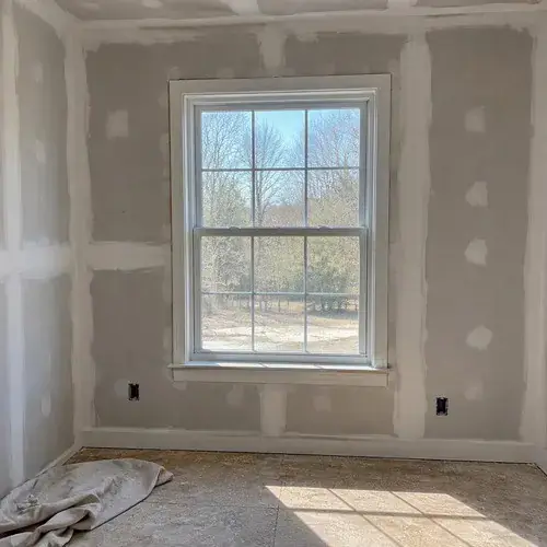 White double-hung window with grids in an unfinished room with taped drywall and plywood